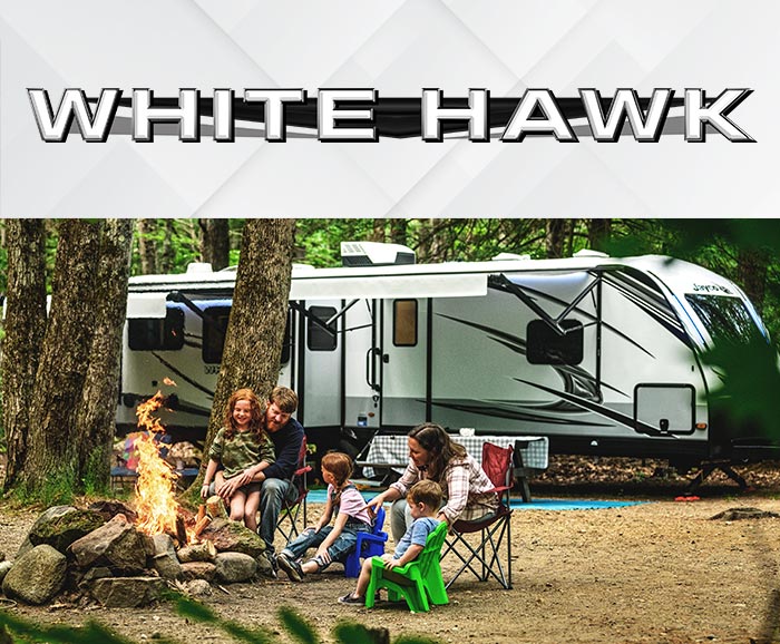 Photo of family sitting around campfire with White Hawk travel trailer in background, and logo above.